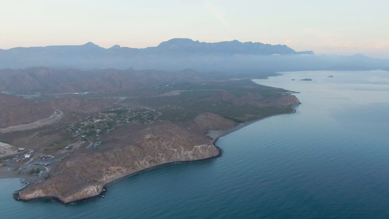 Opening aerial frame over Danzante Bay Loreto, Mexico, with blue water below, a rocky sandy point, a small coastal town, hazy mountains on the horizon, and no visible people.