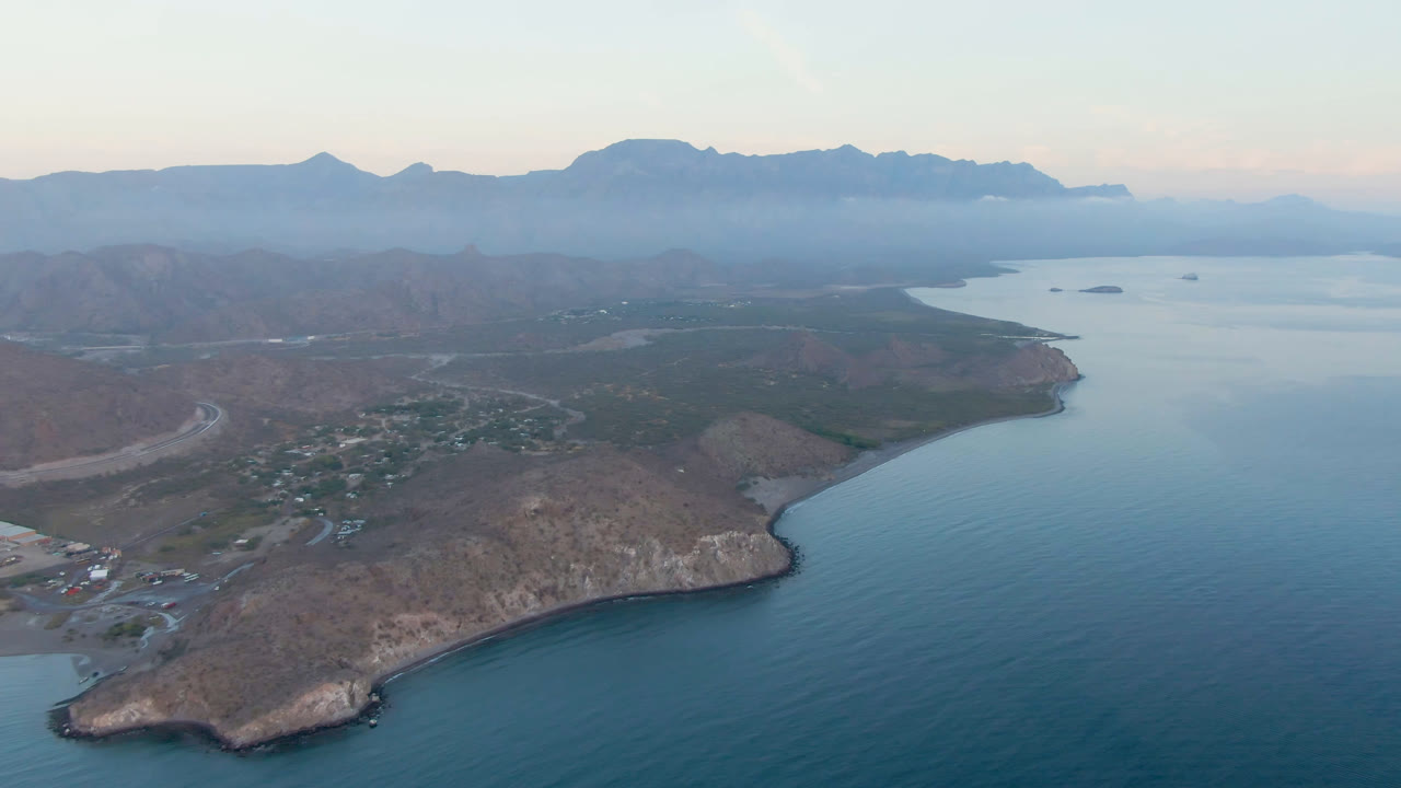 Drone view of a coastal point beside blue water with a small town and mountains in haze.