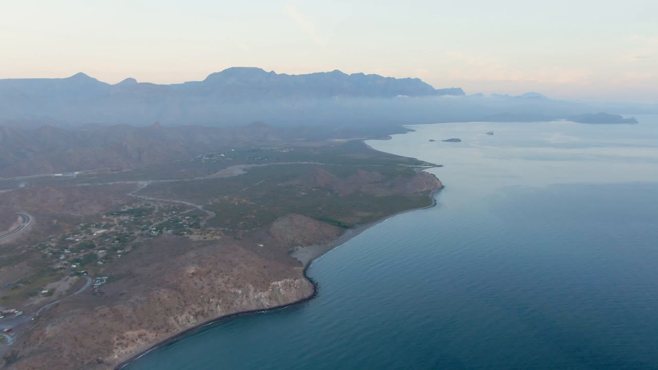 Aerial frame over Danzante Bay in Loreto, Baja California Sur, with blue water on the right, rocky shoreline below, a small town at left, hazy mountains on the horizon, and no visible people.