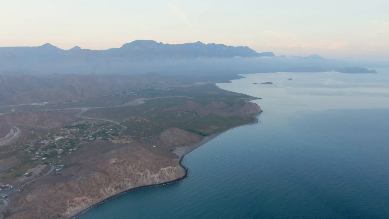Aerial view of Danzante Bay in Loreto, Baja California Sur, with the curving rocky coast lower center, town structures at left, open blue water to the right, distant islands or headlands, and no visible people.