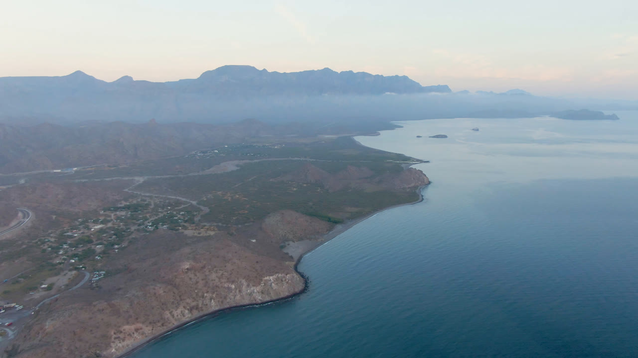 Wide aerial over calm water, rocky coast, small town, and hazy mountain ridge