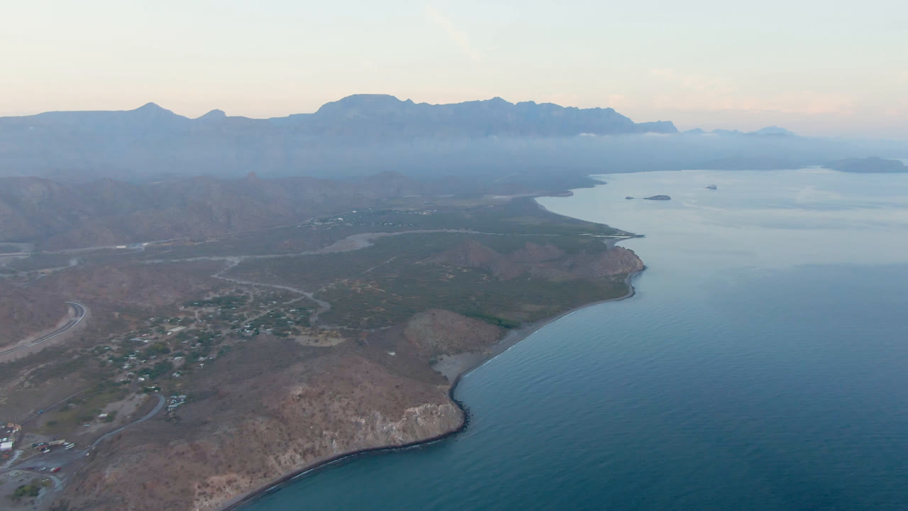 Aerial view one second into the Danzante Bay Loreto clip, showing the coast curving along blue water, clustered town structures and roads on dry land, layered mountains beyond, and no visible people.