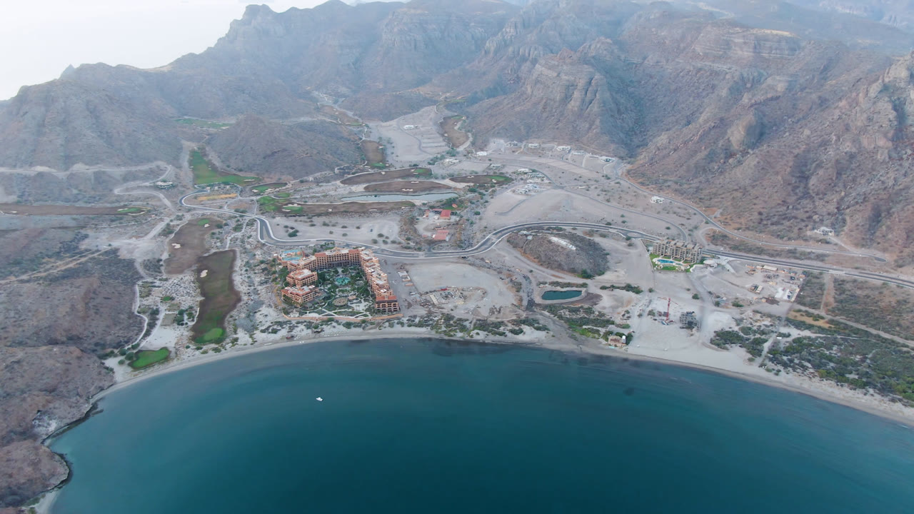 Wide aerial view of bay water, curved shoreline, buildings, roads, and rugged mountains.
