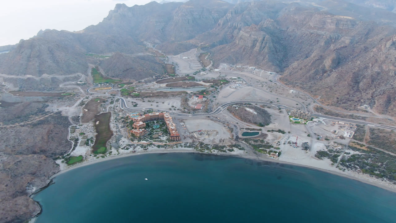 Aerial view of bay water, curved beach, roads, buildings, and rocky mountains.