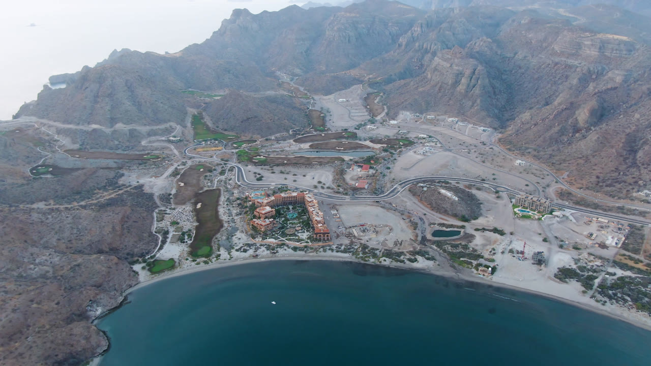 Opening aerial frame over Danzante Bay Loreto, with dark water at the bottom, a curved pale beach, red-roofed buildings, winding roads, arid mountains, and no visible people.