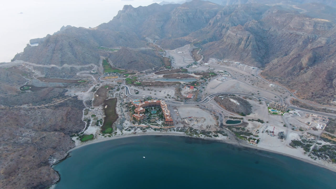 Second aerial frame over Danzante Bay Loreto, showing the waterline lower in frame, a resort-like building cluster, roads crossing pale ground, small water features, mountains, and no visible people.