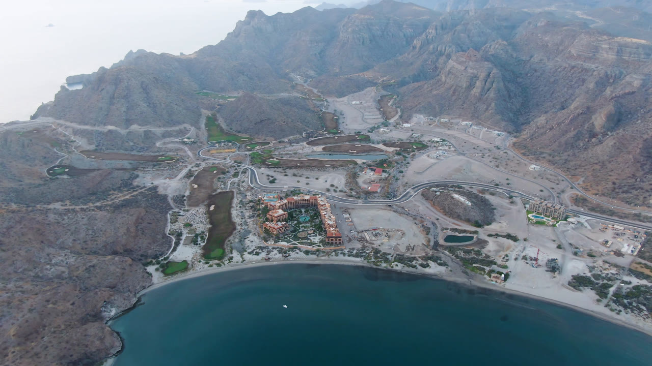 Drone view across a blue bay toward a developed shoreline and brown mountain range.