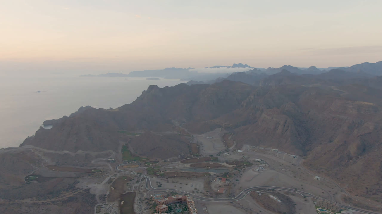 Opening aerial frame over Danzante Bay Loreto, Baja California Sur, with hazy water on the left, mountain ridges across the middle, roads and buildings below, and no visible people.