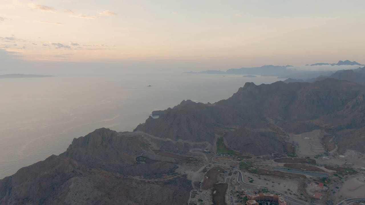 Opening frame over Danzante Bay Loreto, with dark mountain ridges, a winding road below, calm water to the left, pale evening sky, distant islands, and no visible people.