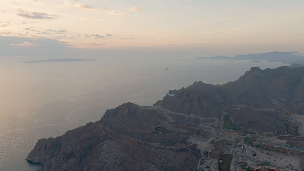 Aerial frame over Danzante Bay Loreto, Baja California Sur, with rugged mountains below, a winding road near the coast, distant islands on the horizon, and no visible people.