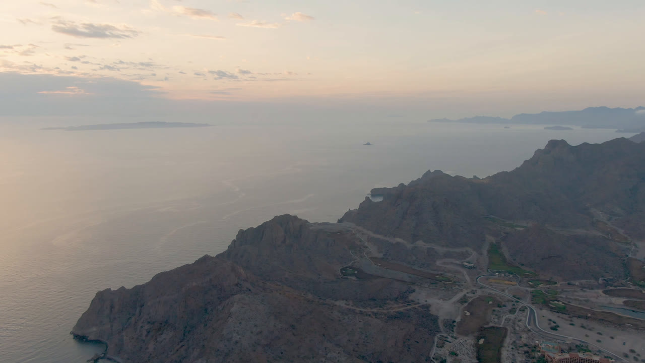 Wide aerial view of Danzante Bay Loreto, Baja California Sur, showing rocky ridges across the lower frame, road cuts through the terrain, calm water beyond, and no visible people.