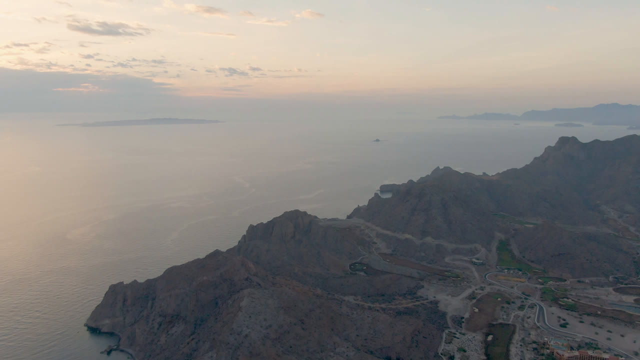 High aerial frame from Danzante Bay Loreto, Baja California Sur, with coastal mountains in shadow, pale ocean water across the middle distance, distant landforms, and no visible people.