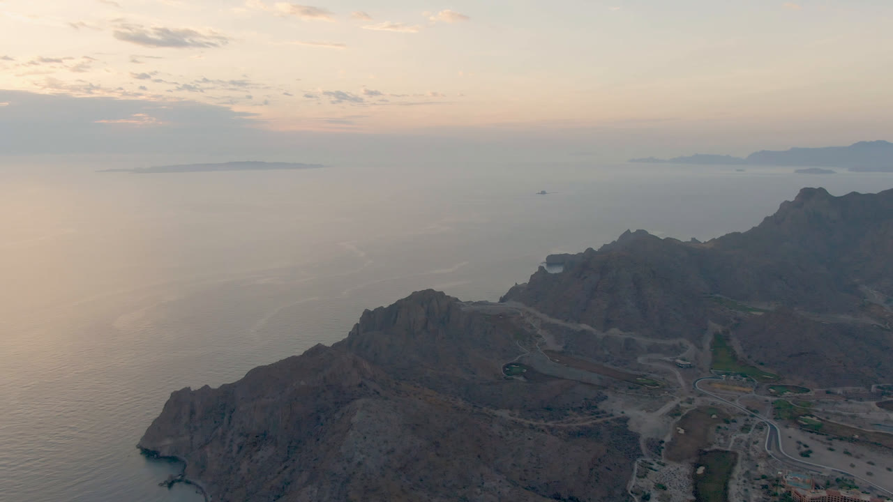 Aerial view in Danzante Bay Loreto, Baja California Sur, with ocean filling the left side, mountain slopes and roads below, distant islands under a warm sky, and no visible people.