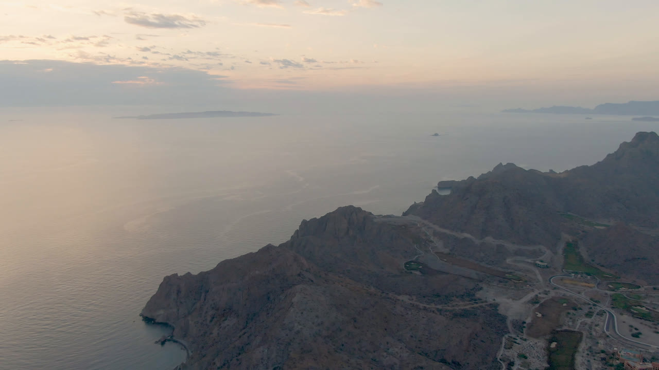 Opening aerial frame over Loreto, Mexico, with rugged cliffs in the lower frame, calm ocean beyond, distant islands on the horizon, and no visible people.