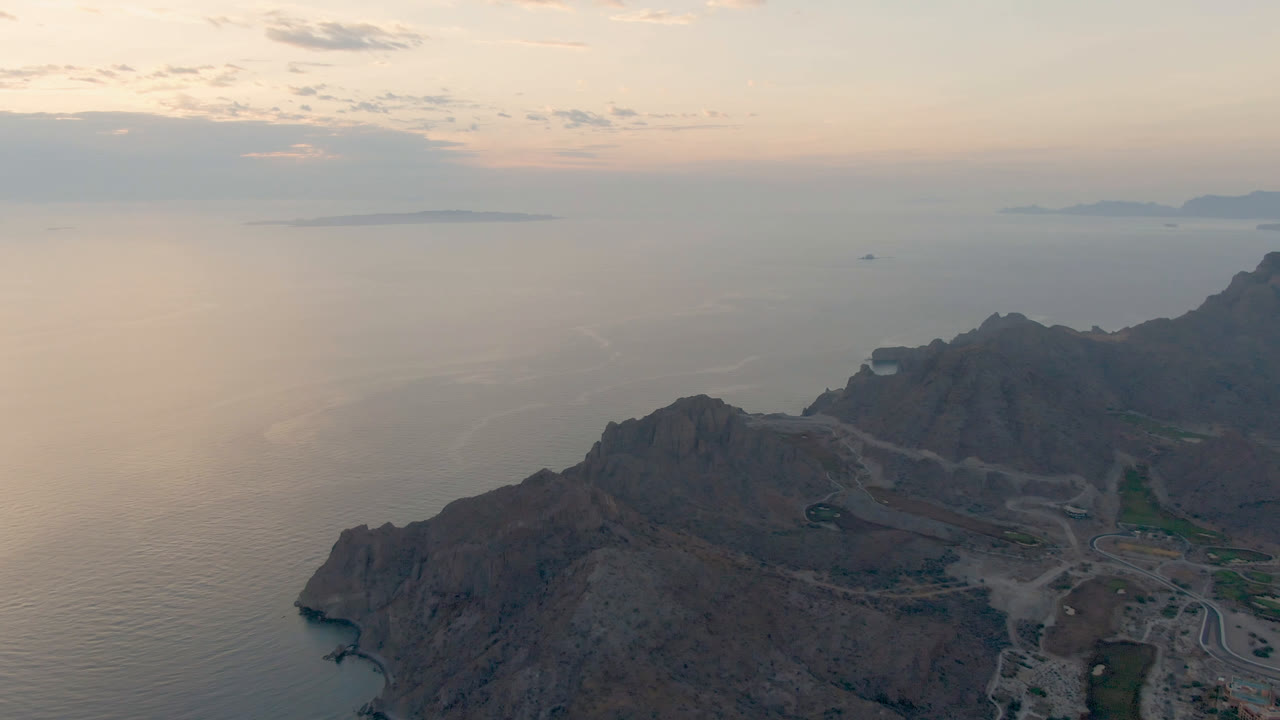 Aerial view in Loreto, Mexico, showing the cliff ridge slightly more centered, open water behind it, low island shapes near the horizon, and no visible people.