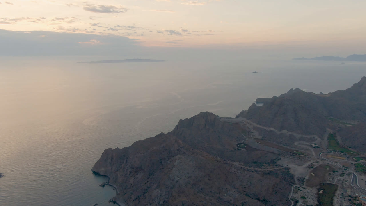 Rocky headland above calm water with distant islands and a muted pink sky.