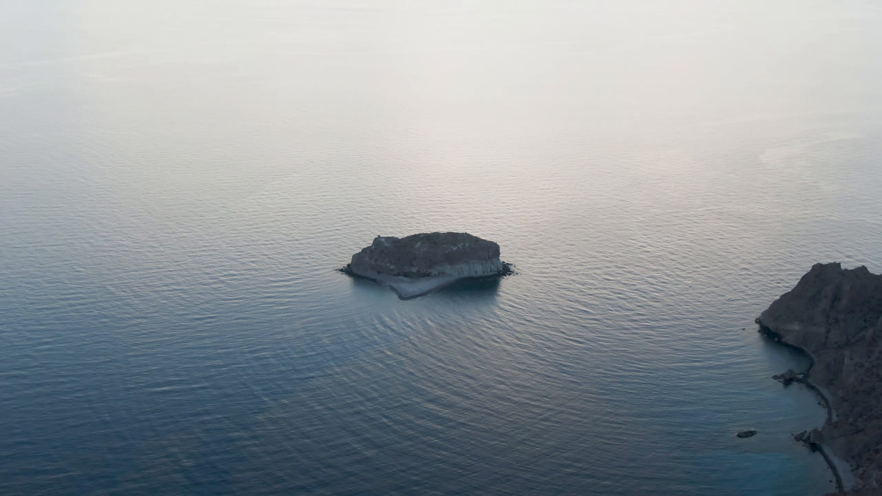 A small rocky island sits left of center in calm blue water at Danzante Bay near Loreto, with hazy daylight and no visible people.