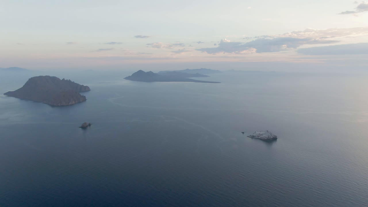 Aerial view over Danzante Bay Loreto with a dark island at left, distant island shapes on the horizon, small boats and a larger white vessel on calm water, and no visible people.