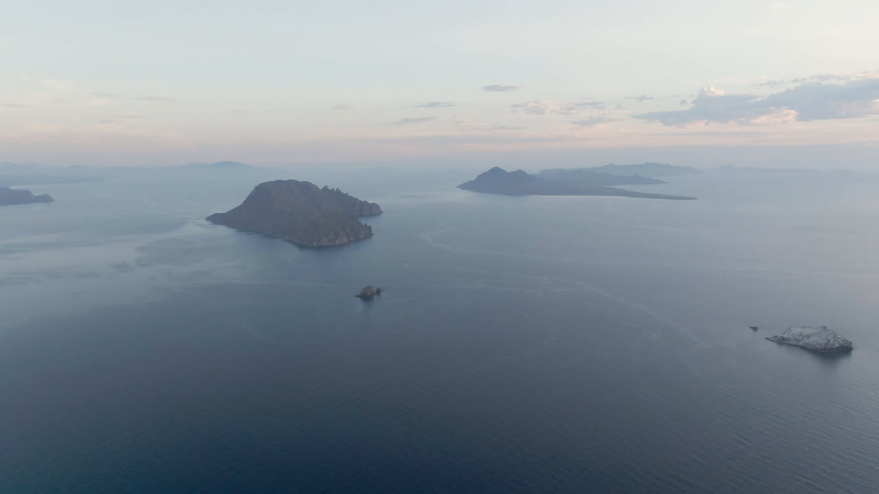 Opening aerial frame over Danzante Bay, Loreto, with a dark island left of center, distant island forms on the horizon, a larger vessel at lower right, and no visible people.