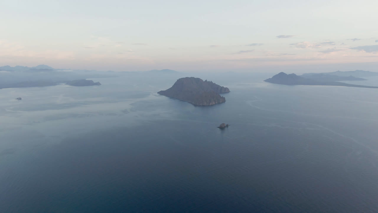 Aerial view of Danzante Bay with the small boat lower in frame, a rounded island left of center, layered island shapes in the distance, soft cloud cover, and no visible people.