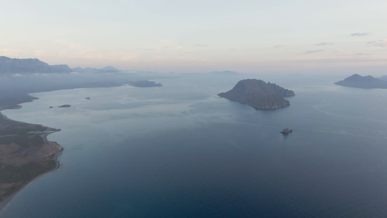 Aerial view over Danzante Bay showing a rounded island near center, a small boat on the water below it, distant landforms, soft sky, and no visible people.