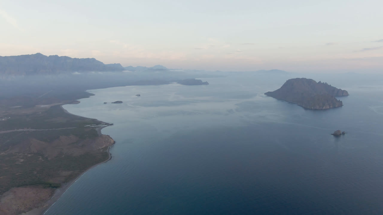 Aerial frame over Danzante Bay in Loreto, Baja California Sur, with a curved foreground shoreline, blue-green water, a rounded island at right, hazy mountain horizon, pale sky, and no visible people.
