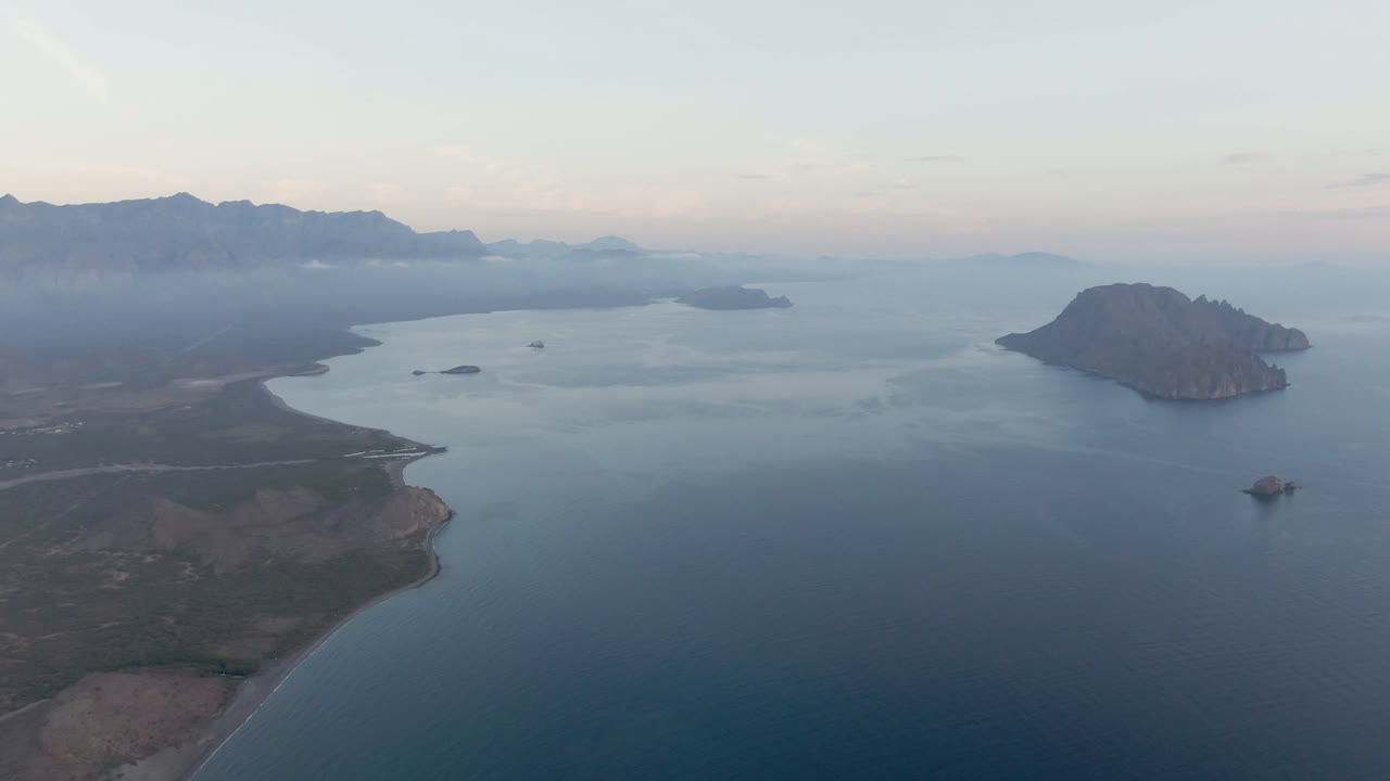 Aerial view of Danzante Bay with the foreground peninsula lower in frame, calm blue water filling the center, small offshore rocks, a rounded island to the right, distant mountains, and no visible people.