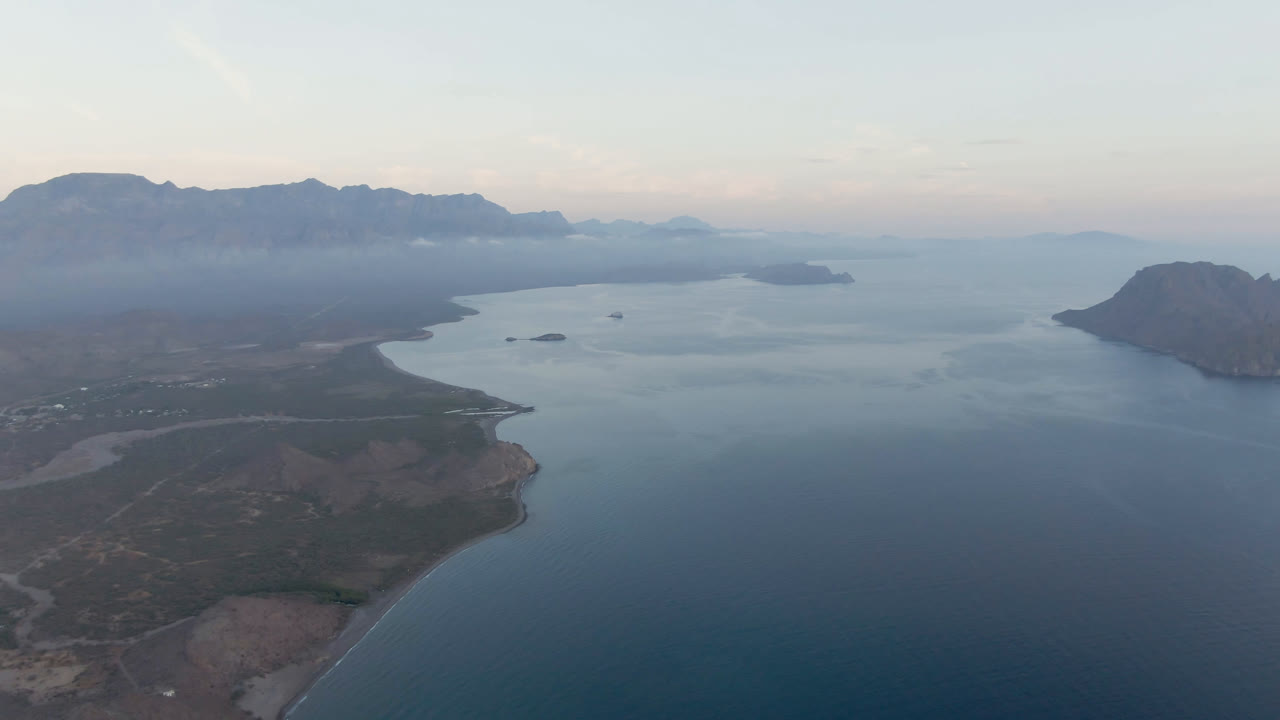 Opening aerial frame over Danzante Bay, Loreto, Baja California Sur, showing a curved shoreline, broad blue water, hazy mountains, pale sky, and no visible people.