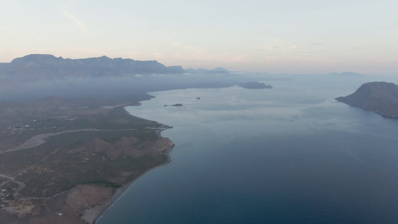 Second aerial frame over Danzante Bay, Loreto, Baja California Sur, with the foreground coast slightly farther across the view, open water at center, hazy mountain ridges, and no visible people.