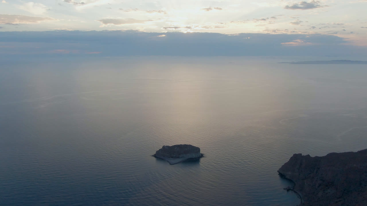 Wide aerial view over Danzante Bay in Loreto, Baja California Sur, with a small rock island low in frame, distant land on the horizon, and no visible people.