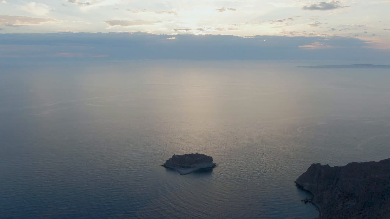 Aerial frame from Danzante Bay showing the rock island slightly left of center, a dark coastline entering from the lower right, pale cloud reflections, and no visible people.