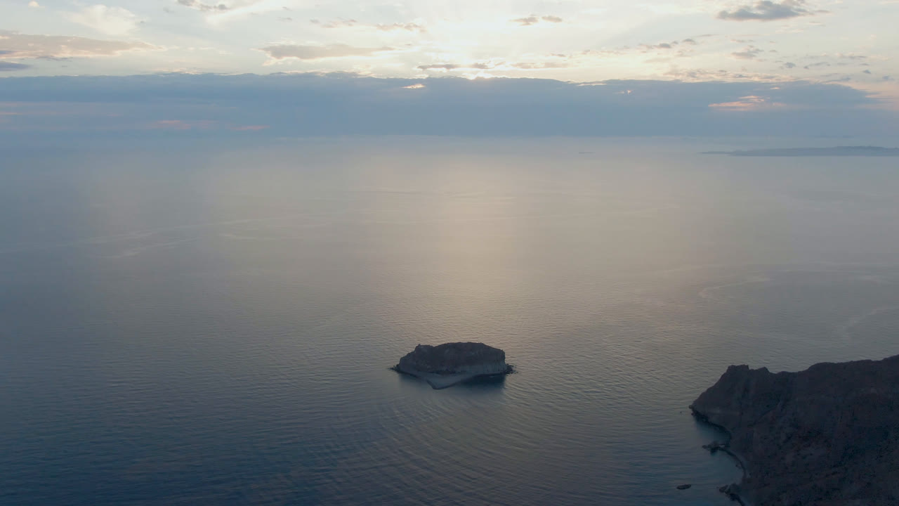Opening frame of Danzante Bay, Loreto showing a small island in calm blue-gray water, rocky coastline at the lower right, distant land on the horizon, cloudy sky, and no visible people.