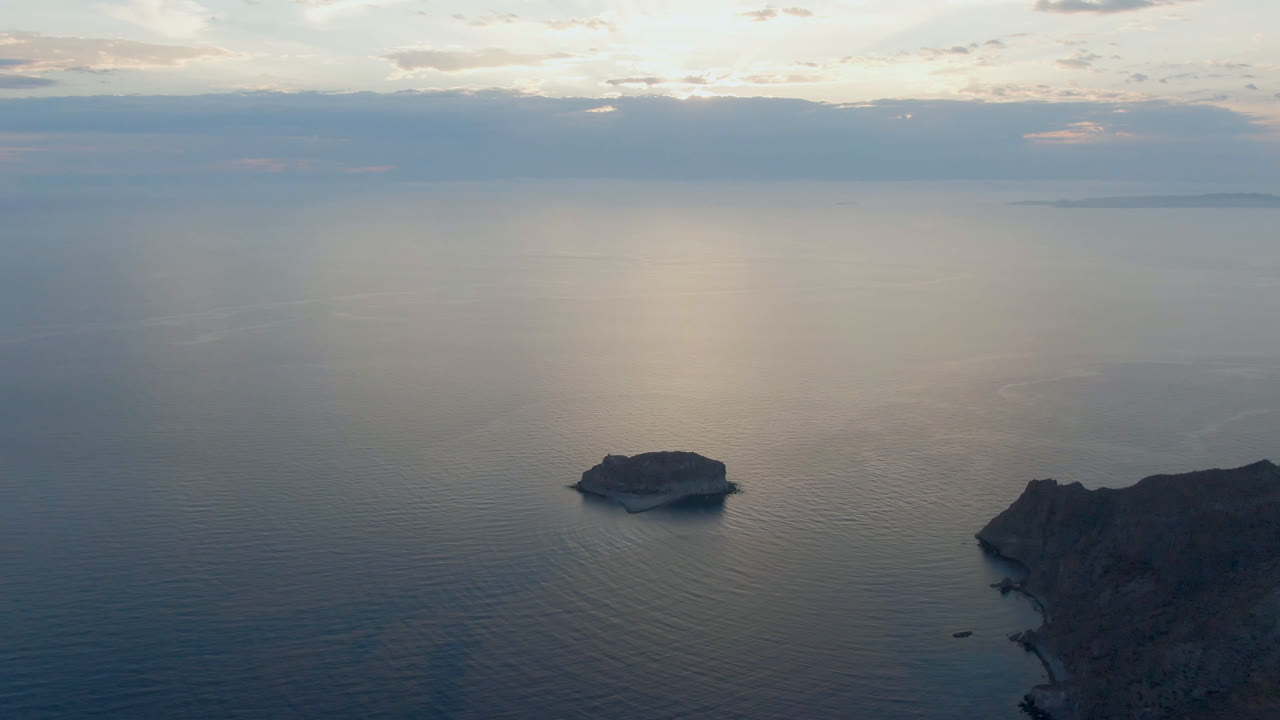 Aerial view over Danzante Bay, Loreto, with a small island left of center, rocky coastline at the right edge, layered clouds on the horizon, and no visible people.
