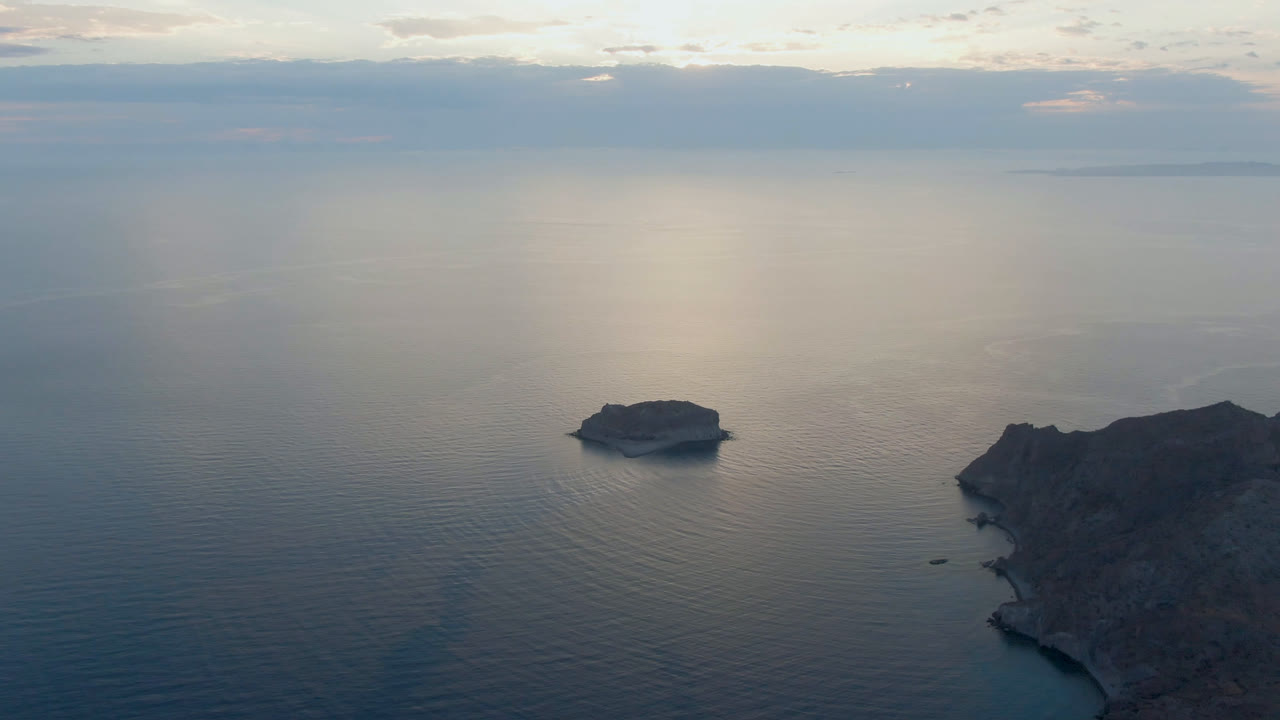 Aerial view over Danzante Bay, Loreto, with a small island left of center, rocky coastline at right, distant horizon islands, cloud reflections on calm water, and no visible people.