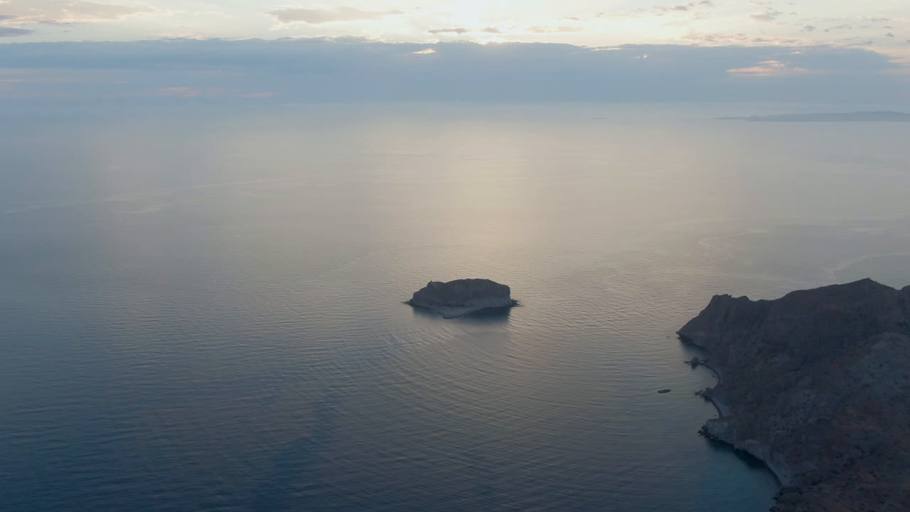 Wide aerial frame from Danzante Bay, Loreto, showing the small island in open water, the rocky landmass along the right edge, muted cloud cover, soft water reflection, and no visible people.