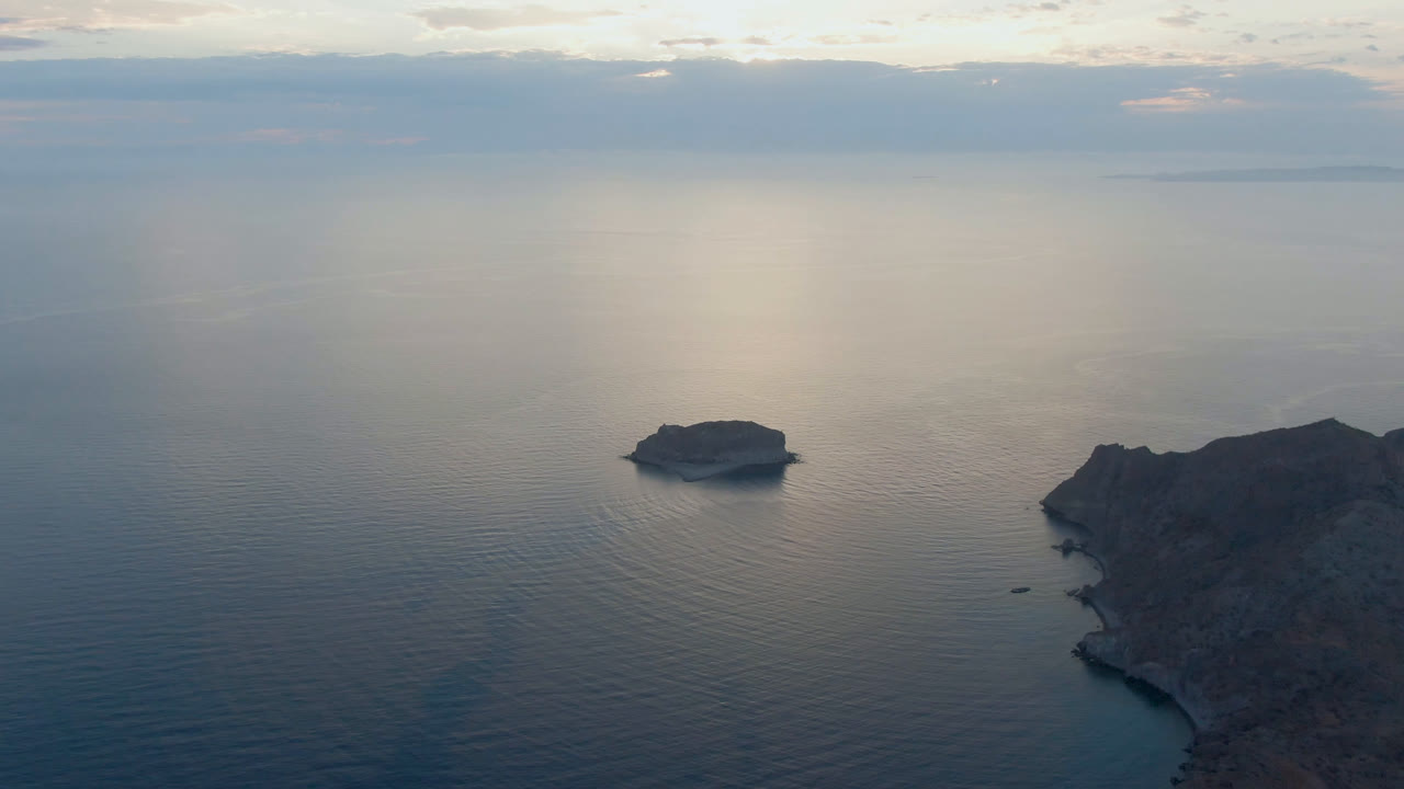 Aerial view in Danzante Bay, Loreto, where the small island sits left of the rocky coastline, pale reflected light spreads across the water, distant land lies on the horizon, and no visible people appear.