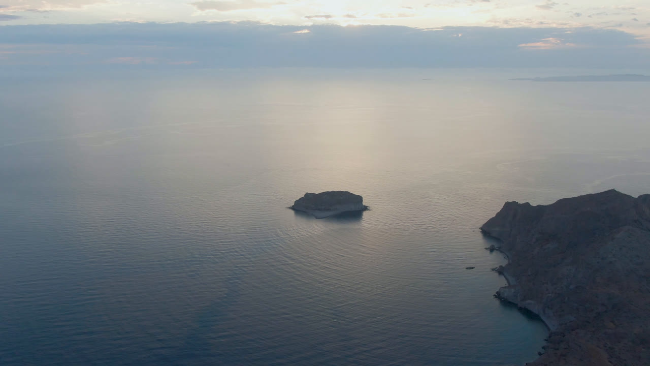 Aerial frame over Danzante Bay, Loreto, with the small island left of center, the rocky shoreline lower right, faint ripples across blue water, cloud bands above, and no visible people.