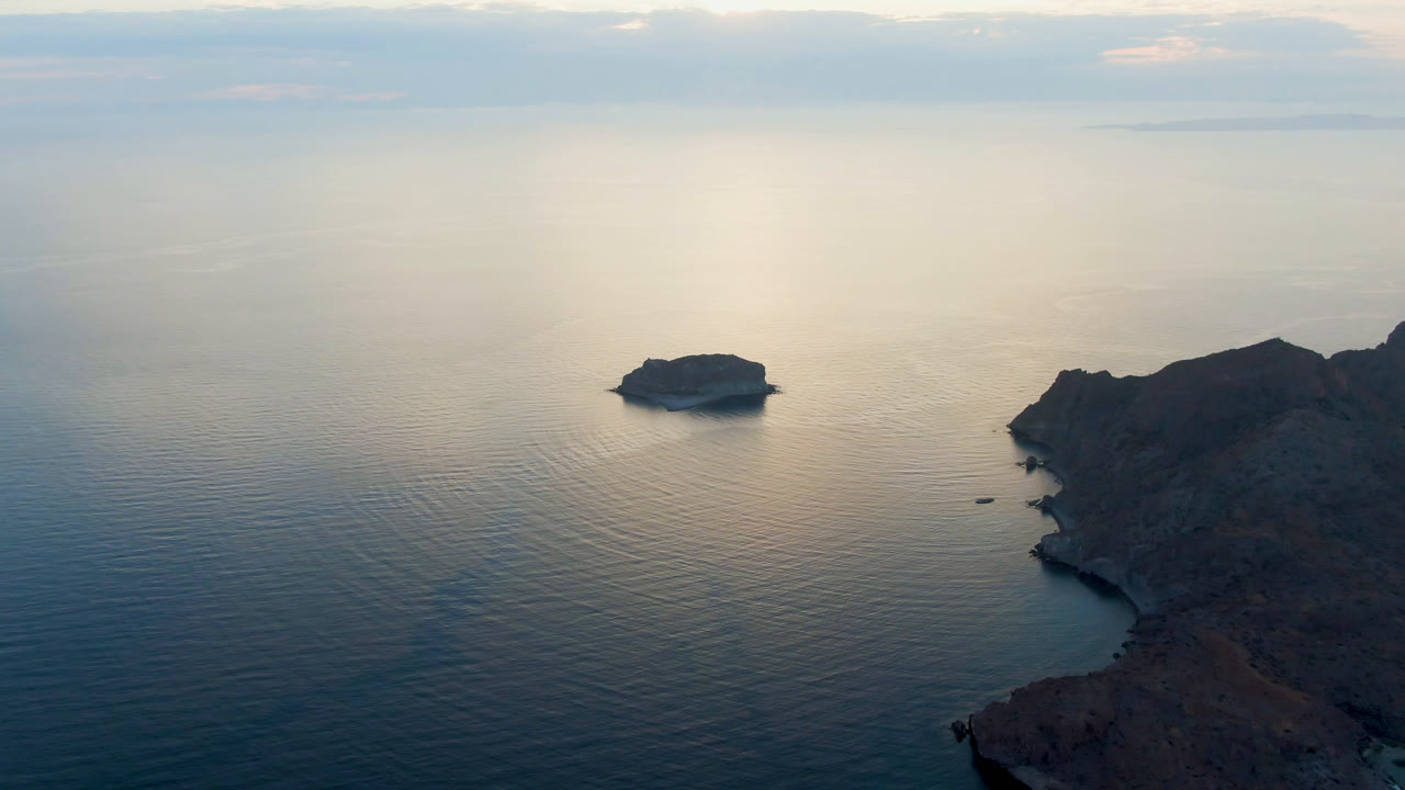 Aerial frame over Danzante Bay Loreto, Baja California Sur, with a small rocky island left of center, dark coastline on the right, soft water reflection, and no visible people.