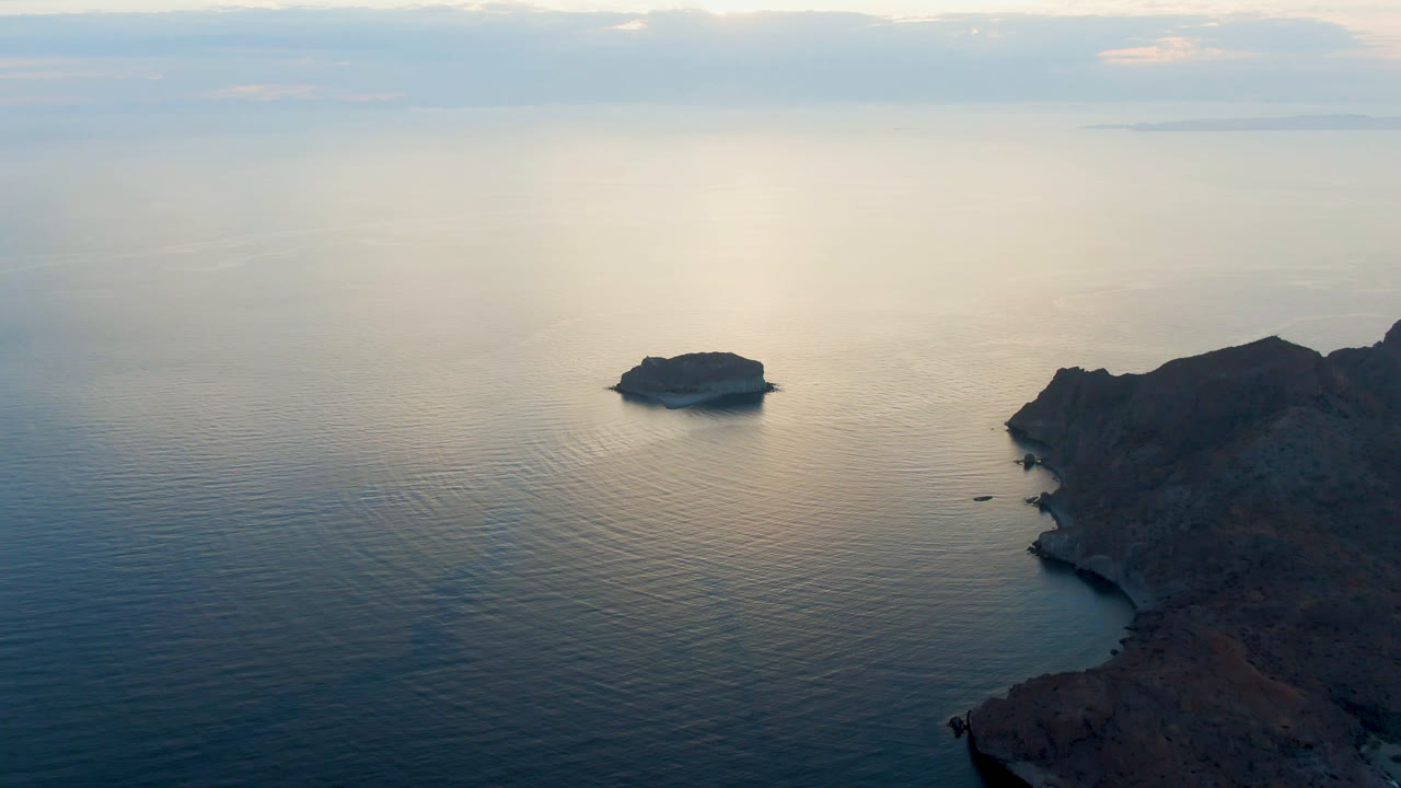At 0:01, Danzante Bay Loreto appears from above with the small island slightly left of center, smooth blue water below, hazy horizon light, and no visible people.