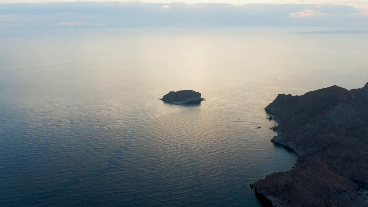 Aerial view of Danzante Bay, Loreto, with a small offshore island left of center, rocky coast at right, reflective ocean water, and no visible people.