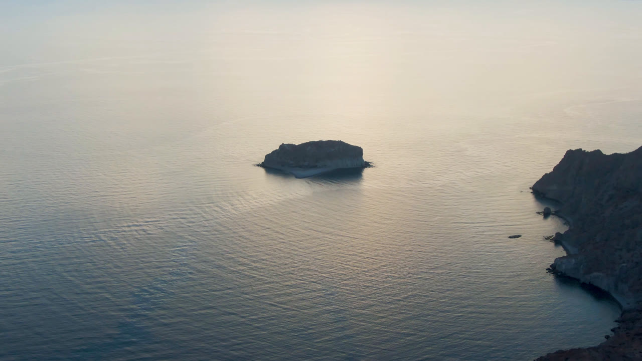 Aerial view over Danzante Bay, Loreto, with a small rocky island left of center, calm reflective water, a dark coastal headland on the right, and no visible people.