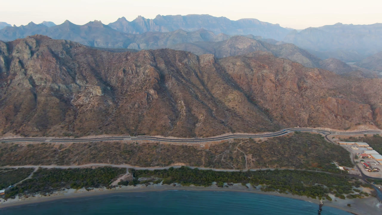 Aerial frame from Danzante Bay Loreto showing blue water, a thin beach, coastal greenery, a road, and steep brown mountains, with no visible people.