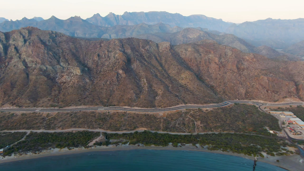 Wide aerial frame at Danzante Bay Loreto showing turquoise water, shoreline brush, a road along the mountain base, low coastal buildings, and no visible people.