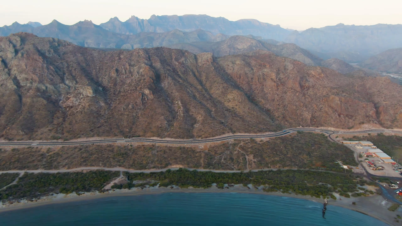 Aerial view of blue bay water, shoreline, road, and rugged mountains.