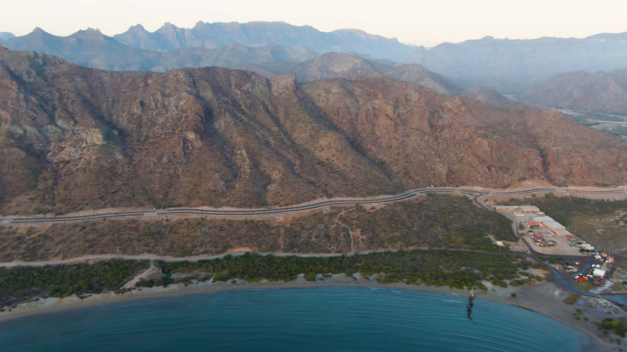 Aerial view of bay water, shoreline, road, and rugged mountains.