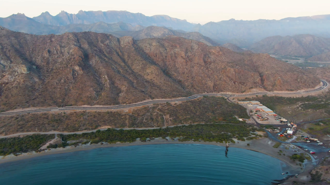 Opening aerial frame over Danzante Bay Loreto, Baja California Sur, with blue water in the foreground, a pale shoreline road, layered brown mountains, and no visible people.