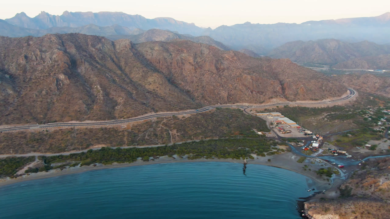 Opening aerial frame over Danzante Bay in Loreto, Baja California Sur, with blue water in front, a narrow beach, a curving road, brown mountains, and no visible people.