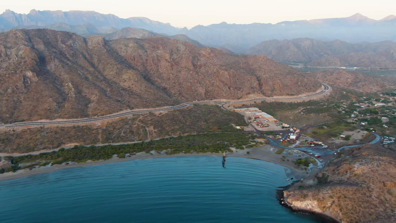 Aerial view of blue bay water, shoreline trees, road, buildings, and brown mountains.