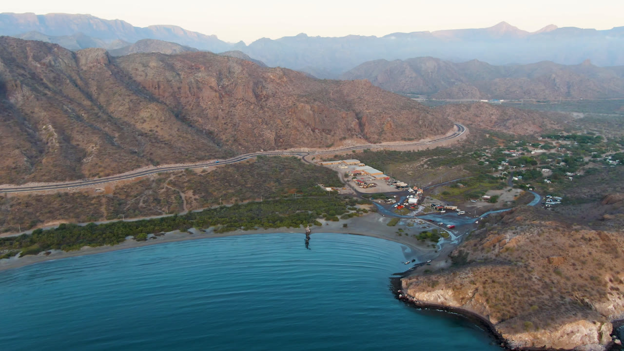 Wide aerial view of blue water, rocky shoreline, road, buildings, and mountains.