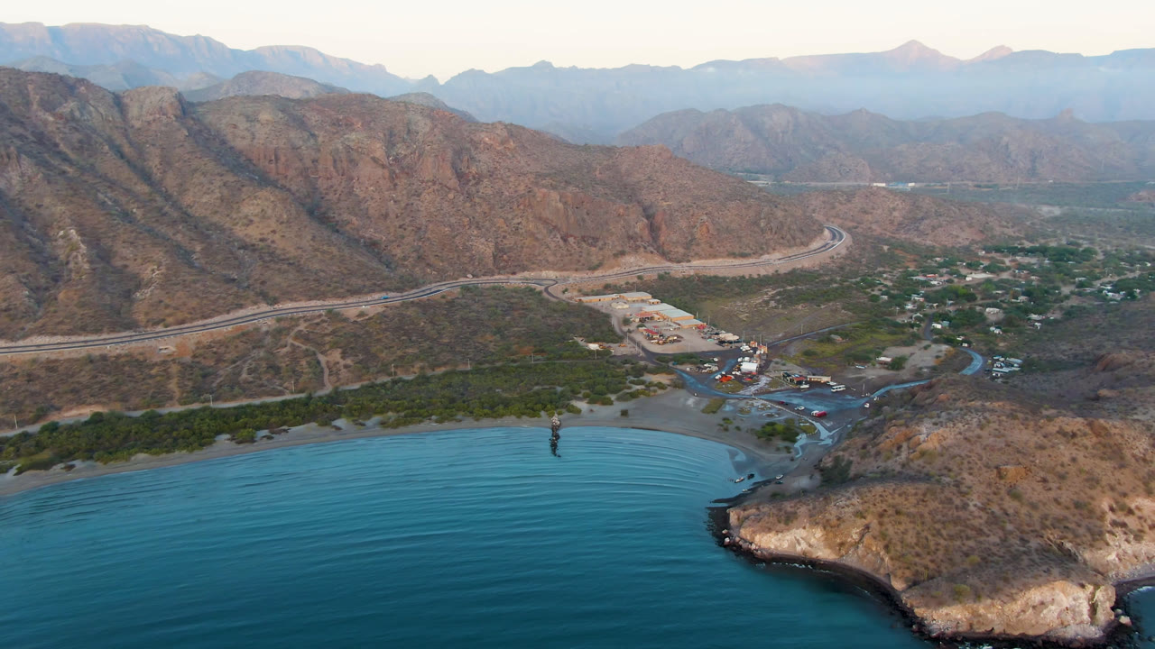 Aerial view of Danzante Bay Loreto with calm blue water across the lower frame, a narrow shoreline, clustered buildings near a road, rugged mountains, and no visible people.